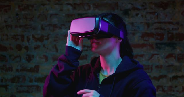 Woman wearing VR headset enjoying immersive gaming experience in dimly lit room. Pink and blue neon lights casting ambient glow. Ideal for tech articles, virtual reality promotional materials, gaming blogs, and futuristic technology concepts.