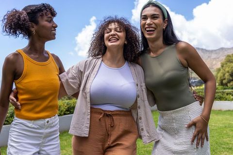 Three diverse women laughing together in sunlit park hugging arms casual summer fashion
