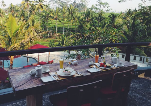 Outdoor breakfast setting featuring wooden table with plates of food and drinks, overlooking lush tropical rice fields and a pool. Ideal for use in travel blogs, tourism promotions, resort marketing, and healthy eating advertisements.