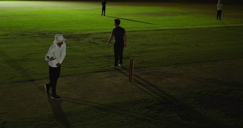 Night cricket practice: young player standing by stumps under floodlights on grass pitch