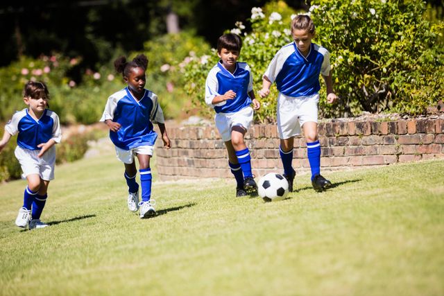 Children in blue and white soccer uniforms are playing a match in a park. They are running on a grassy field, focused on the soccer ball. This image can be used for promoting youth sports programs, outdoor activities, teamwork, and healthy lifestyles for children.