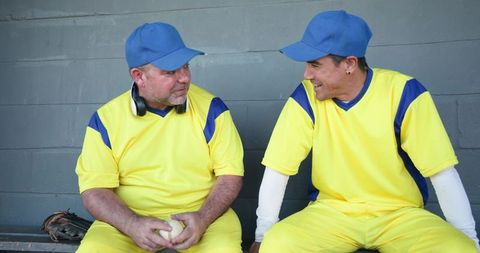 Two mature baseball teammates chatting on dugout bench in yellow blue uniforms