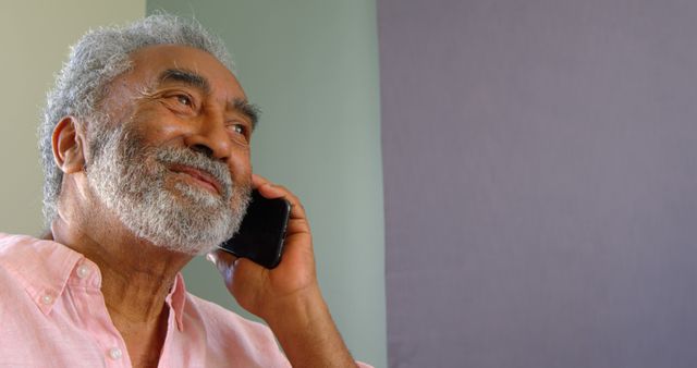 Senior man with gray hair and beard wearing casual shirt talking on smartphone, looking happy. Useful for topics related to technology use by seniors, communication, happiness, or casual conversations.