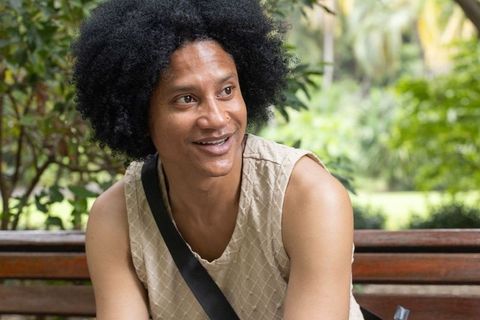 Relaxed african american man smiling on park bench wearing beige top, crossbody bag strap
