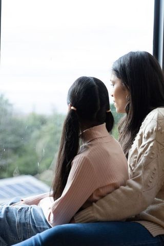 Asian mother and daughter sharing quiet window moment with solar panels in cozy knitwear