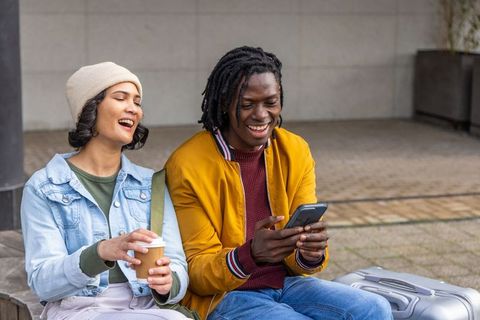 Diverse couple laughing and checking smartphone on urban bench with coffee and luggage