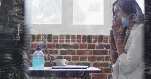Woman adjusting face mask while checking laptop with health alert icons at office desk