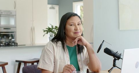 Mature asian woman podcasting in modern open-plan kitchen workspace with laptop and mic