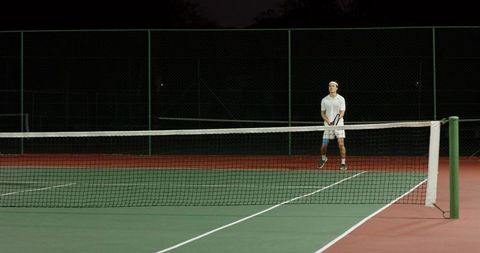 Night tennis player standing on empty hardcourt holding racket in white polo and headband