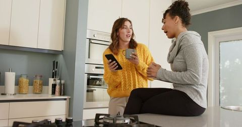 Sharing morning coffee and chatting in modern kitchen, friends using phone, cozy sweaters