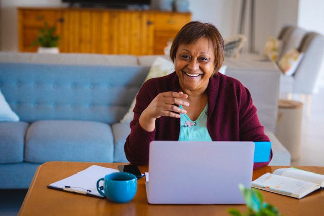 Senior African American woman smiling while using a laptop at home. Ideal for illustrating concepts related to retirement lifestyle, home finances, communication, and the use of wireless technology by elderly individuals. Suitable for articles, blogs, and advertisements focusing on senior citizens, remote work, and digital literacy.