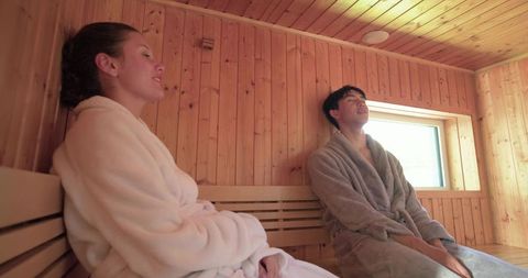Diverse couple relaxing in cedar sauna wearing plush bathrobes, serene steamroom retreat