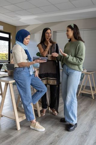 Diverse female team collaborating in modern office, discussing project with tablet and mug