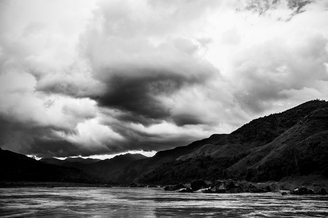 The black and white photograph of a mountain river with dramatic storm clouds above captures an intense and moody atmosphere. Ideal for use in projects that want to convey natural beauty, season changes, and an adventurous or serene theme. Can be used in travel blogs, nature documentaries, desktop backgrounds, and scenic artwork.