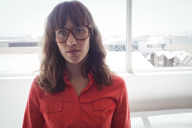 This image shows a serious female designer standing by a window in an office. She is wearing glasses and a red shirt, giving a professional and thoughtful look. The natural light from the window enhances the modern and creative atmosphere. This image can be used for articles or advertisements related to professional women, creative industries, office environments, and business settings.