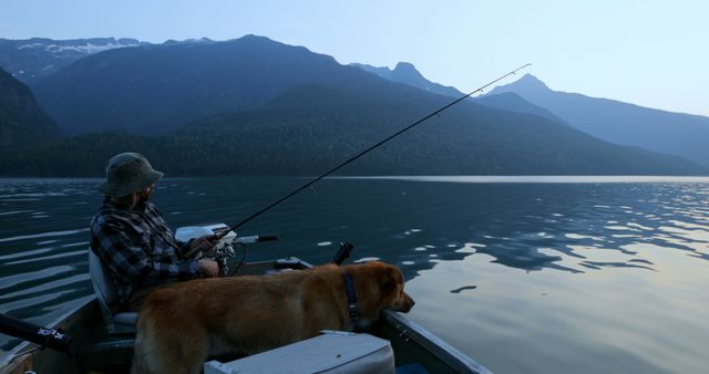 A middle-aged Caucasian man enjoys fishing on a serene lake with his loyal dog by his side, with copy space. Surrounded by majestic mountains, the scene captures a moment of tranquility and companionship in nature.