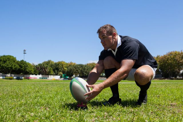 Rugby player holding ball while kneeling on grassy field against clear sky