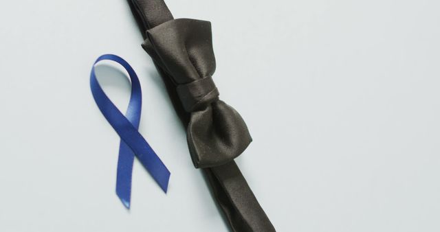 Blue awareness ribbon placed next to black bow tie on light color background. Perfect for campaigns promoting prostate cancer awareness, formal wear advertisements, and health-focused initiatives.