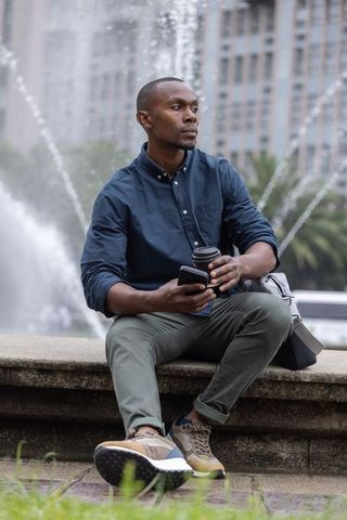 Urban professional sitting by fountain holding coffee and phone wearing smart-casual outfit