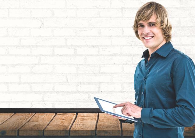 Man in blue shirt using a tablet while smiling, standing against a white brick wall. Ideal for concepts related to modern technology, digital communication, business presentations, and office environments.