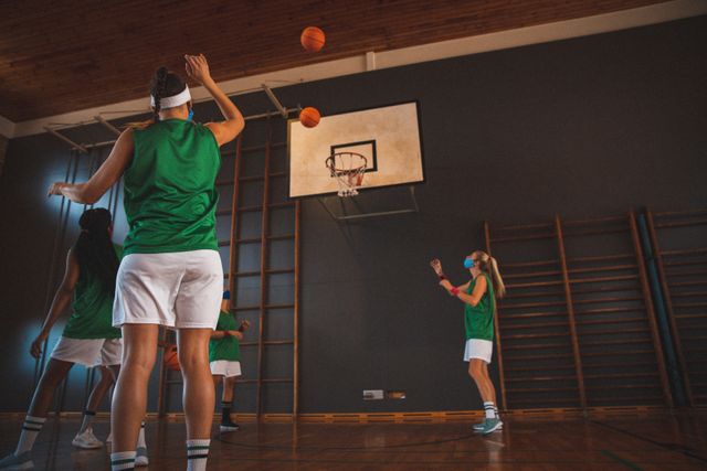Diverse female basketball team wearing face masks practicing shooting hoops in court. basketball, team sports training at an indoor court during covid 19 coronavirus pandemic.
