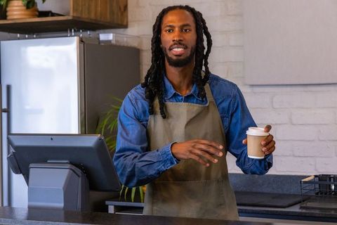 Urban barista smiling and serving takeaway coffee at cafe counter with touchscreen pos and apron