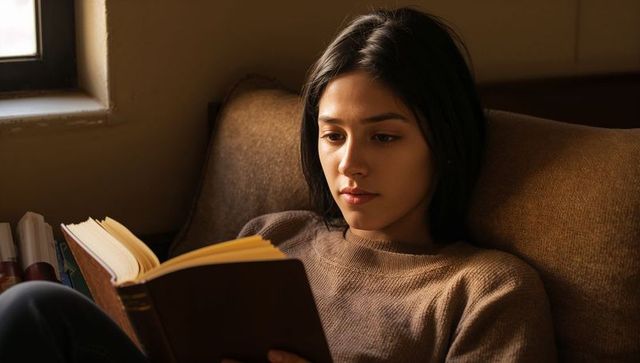 Young woman reading on couch by window in cozy brown sweater, sunlit quiet moment, solitude