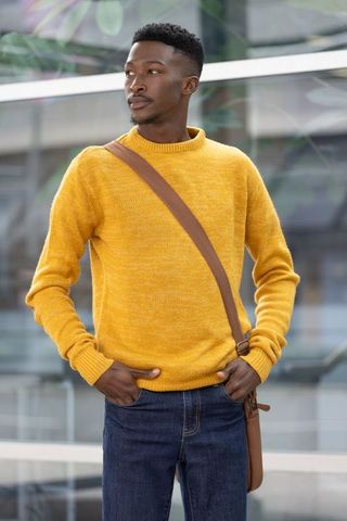 African american man standing at glass facade wearing mustard sweater and crossbody bag