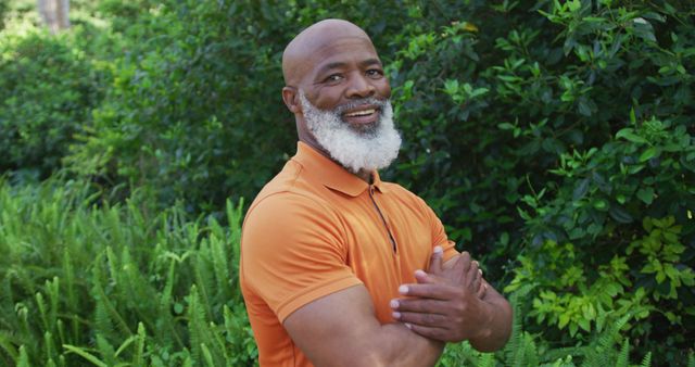 Mature man with a white beard smiling confidently while standing outdoors surrounded by lush greenery. He wears an orange polo shirt and has arms confidently crossed. This image can be used for promoting active senior lifestyles, nature-related themes, health and wellness content, or positive living campaigns. Useful for websites, social media posts, and advertisements targeting mature audiences.