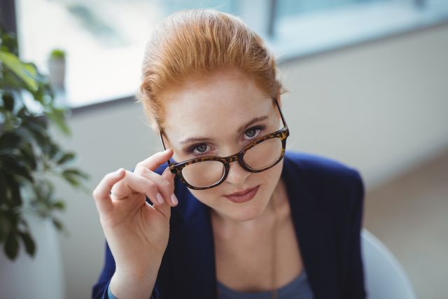 Businesswoman adjusting glasses while looking at camera in modern office. Ideal for corporate websites, business presentations, career-related articles, and professional development materials.