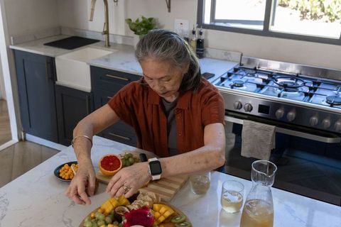 Senior chinese woman arranging vibrant fruit platter on kitchen island wearing smartwatch