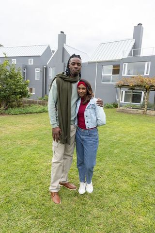 Multicultural couple posing on green lawn in front of modern grey townhouses