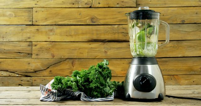 A blender filled with ingredients sits ready for making a smoothie, with a bunch of fresh kale and a cloth napkin on a wooden surface, with copy space. Preparing nutritious green smoothies is a popular way to maintain a healthy diet.