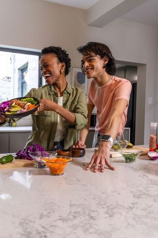 Diverse couple preparing vibrant rainbow salad on marble island, sharing joyful cooking moment
