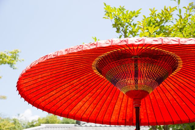 Traditional red Japanese umbrella providing shade on a sunny day with a clear blue sky. Perfect for themes related to Japanese culture, outdoor décor, summer scenes, and traditional craftsmanship. Ideal for travel blogs, cultural articles, seasonal promotions, and decorative inspiration.