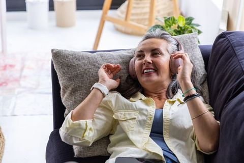 Relaxed middle-aged woman listening to music on sofa with pink wireless headphones