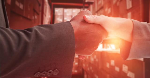 This image shows a close-up of two business people shaking hands, symbolizing cooperation and sealing a professional deal. The background, filled with storage and shipping boxes, indicates a warehouse environment. This can be useful for themes like business partnerships, logistics, agreements, teamwork, and diversity in industry settings.