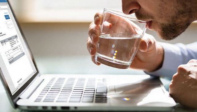 Businessman drinking water while reviewing document on laptop, focused desk productivity