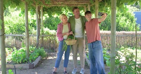 Family taking selfie in backyard vegetable garden under vine-covered pergola during harvest