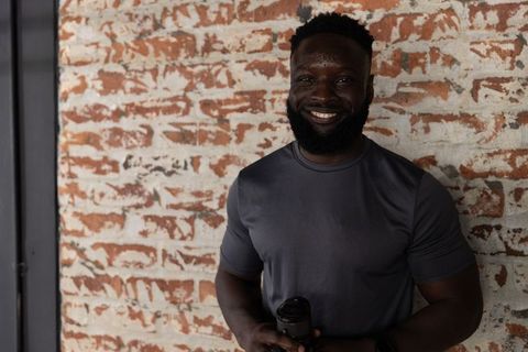 African american man smiling holding black mug by distressed brick wall, fitness portrait