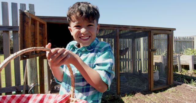 Young boy holding a picnic basket with a big smile near a chicken coop on a sunny day. This image can illustrate family farming, childhood joy, outdoor activities, and rural life. Suitable for use in educational articles, promotions for outdoor events, or children's books.