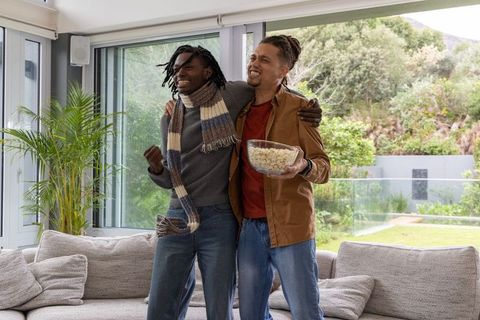 Laughing diverse friends celebrating movie night at home holding popcorn by sunny window
