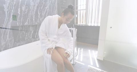 Woman sitting in white bathrobe beside freestanding tub in bright marble spa bathroom