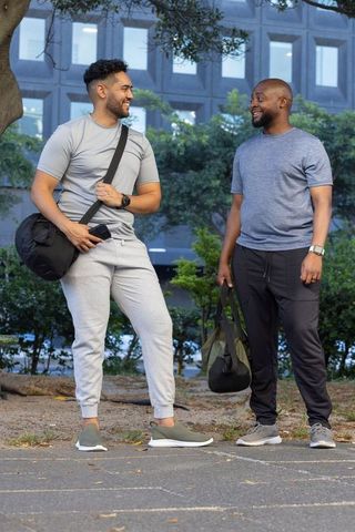 Two men smiling and chatting in workout clothes while holding duffel bags in urban plaza