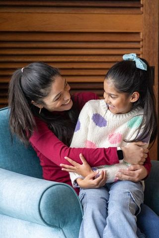 Indian mother and daughter hugging on blue chair smiling, cozy home moment with smartwatch
