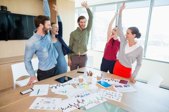 Five business professionals are celebrating with raised hands in a bright, modern office meeting room. They appear excited and happy, standing around a conference table filled with charts, graphs, and office supplies. This image can be used for illustrating corporate success, teamwork, or a productive business environment.