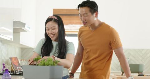 Asian couple planting herbs on sunlit modern kitchen island, smiling, urban indoor gardening
