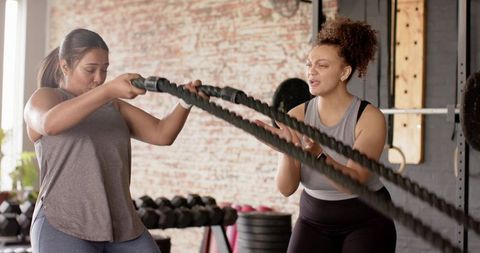 Diverse women doing intense battle rope workout with coach during team training in sunlit gym