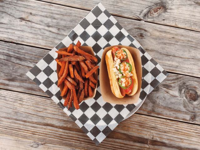 Gourmet hot dog served with a side of sweet potato fries on a wooden table, creating a rustic and appetizing setting ideal for promoting food trucks, casual dining, summer meals, and various comfort food concepts on restaurant menus or lifestyle blogs.