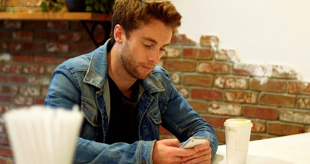 Young man wearing a denim jacket is sitting at a table, engaged in texting on his smartphone. He is in a cafe with a brick wall background. A cup of coffee is on the table nearby. Ideal for concepts related to modern lifestyle, technology, communication, and casual dining.